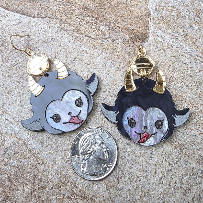May include: A pair of dangling earrings featuring stylized creature faces with black and iridescent white acrylic, gold-tone horns, and red glitter tongues. A US quarter coin is placed between the earrings for scale, showing their relative size.