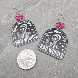 May include: A pair of black and white acrylic earrings featuring a skeleton graphic and the text Good Moms Say BAD WORDS, with pink heart-shaped connectors and silver-toned hooks, displayed next to a United States quarter for scale.