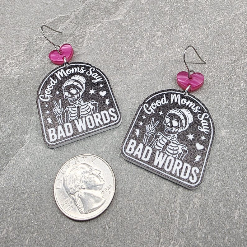 May include: A pair of black and white acrylic earrings featuring a skeleton graphic and the text Good Moms Say BAD WORDS, with pink heart-shaped connectors and silver-toned hooks, displayed next to a United States quarter for scale.