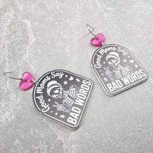 May include: A pair of dangle earrings featuring a black arch-shaped pendant with a white skeleton graphic making a peace sign. The text reads Good Moms Say BAD WORDS. Each earring has a small pink heart-shaped connector and a silver metal hook.