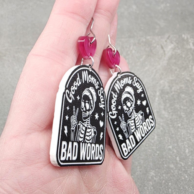 May include: A pair of dangle earrings featuring a black and white arched design with a skeleton making a peace sign. The text reads Good Moms Say BAD WORDS. Each earring is topped with a small, translucent pink heart-shaped acrylic charm.
