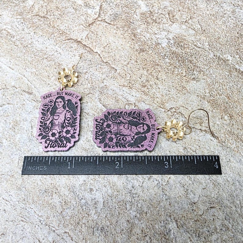 May include: Two lavender-colored rectangular earrings featuring a black graphic illustration of a person surrounded by flowers and a bird, with the text "RAGE... BUT MAKE IT Floral". Each earring has a gold-tone flower-shaped stud and hook.