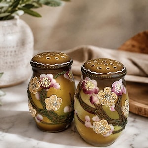 May include: A pair of rounded salt and pepper shakers featuring a gradient olive green and brown finish. Each shaker is decorated with raised, textured floral motifs in shades of pink and gold, with brown branches and a perforated metal lid.