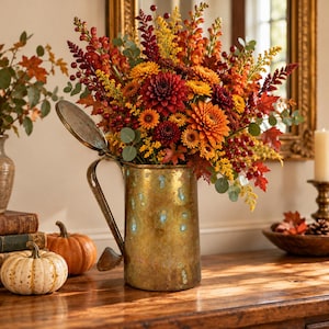 May include: A rustic brass-toned metal pitcher with a hinged lid, filled with an autumnal floral arrangement featuring deep red dahlias, orange chrysanthemums, yellow sprigs, red berries, and green eucalyptus leaves.