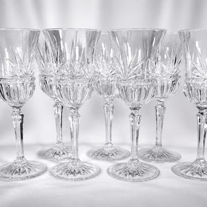 May include: A set of six clear crystal wine glasses. Each glass features a detailed, cut-glass design on the bowl and base, with a slender stem. The glasses are arranged in a row, showcasing their elegant craftsmanship.