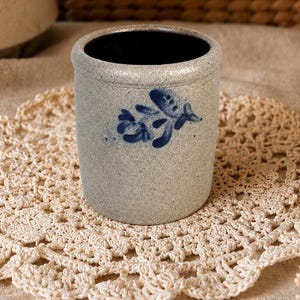 May include: A cylindrical stoneware crock featuring a speckled light gray finish and a hand-painted blue floral motif on the front. The vessel has a thick, rounded rim and a smooth, dark interior, presenting a sturdy, utilitarian design for storage.