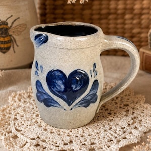 May include: A speckled beige ceramic pitcher featuring a hand-painted dark blue heart design on the front. The pitcher has a curved handle, a pouring spout, and a dark blue interior finish. The exterior surface has a textured, speckled appearance.