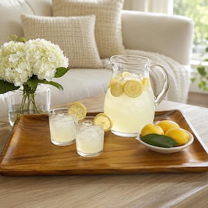 May include: A rectangular wooden serving tray holding a glass pitcher of lemonade with lemon slices and ice, two matching glasses filled with lemonade and ice, and a small white bowl containing three whole yellow lemons with a green leaf.