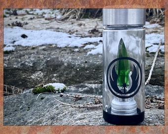 May include: A clear glass water bottle with a silver lid and black base. Inside is a green crystal with a black and white graphic. The bottle is set against a natural, snowy background.