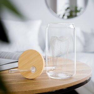 Pode incluir: Um copo de vidro transparente em forma de lata com um design de dente gravado em detalhe. O copo inclui uma tampa de bambu e um canudo transparente. O copo está sobre uma mesa de madeira. O fundo está desfocado, com uma almofada branca e um espelho.