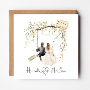 May include: A wedding invitation card with a watercolor illustration of a couple sitting on a swing adorned with white flowers. The text "Mr & Mrs" is on a heart-shaped tag hanging from the swing. The date "14.01.2025" is on a tag hanging from the swing. The text "Hannah and Matthew" is at the bottom of the card.