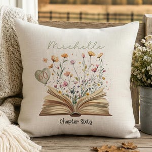 May include: A square off-white throw pillow featuring a printed illustration of an open book with colourful wildflowers emerging from the pages. A heart-shaped balloon with the number 60 is attached, with the name Michelle and Chapter Sixty printed.