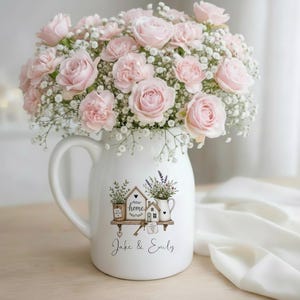 May include: A white ceramic pitcher filled with a large bouquet of light pink roses and white baby's breath. The pitcher features a graphic of a house on a shelf with the text "new home" and "Jake & Emily" printed in black script below.