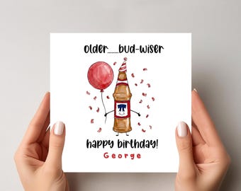 May include: A birthday card held by hands, featuring a cartoon beer bottle with a party hat and a red balloon. The card reads "older...bud-wiser" and "happy birthday! George" in black and red text, with confetti.