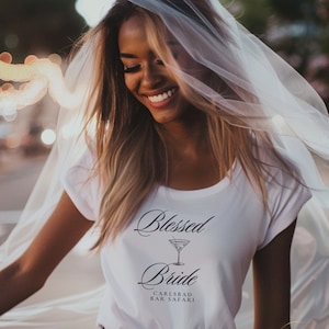 May include: A white t-shirt with the words "Blessed Bride" and a martini glass graphic. The text "Carlsbad Bar Safari" is below the graphic. A sheer veil is draped over the person's head.