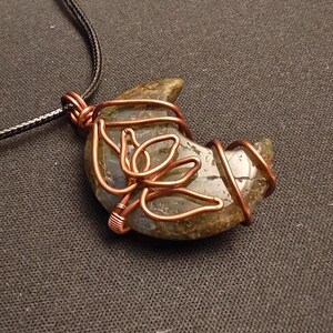 May include: A crescent moon-shaped pendant necklace. The pendant is a stone wrapped in copper wire, with a floral design. The necklace has a black cord. The stone is a mix of green, gray, and brown.