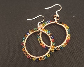 May include: A pair of copper wire hoop earrings with colorful glass beads. The beads are arranged in a rainbow pattern.
