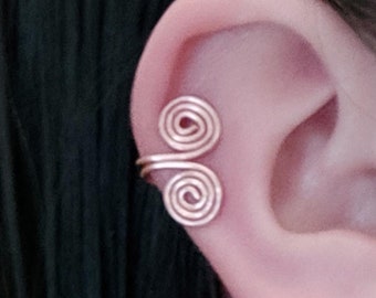 May include: A close-up of a person's ear with a gold ear cuff. The ear cuff is made of two intertwined spirals.