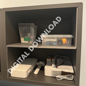 Puede incluir: Una estantería gris con dos estantes. El estante superior tiene una cesta de alambre de malla, un pequeño contenedor de plástico transparente con tapa amarilla y una pequeña caja gris. El estante inferior tiene una caja rectangular blanca, una herramienta blanca y gris, una caja rectangular blanca con un cable blanco y una bolsa de plástico blanca con un cable rojo y verde.