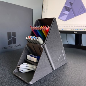Pode incluir: Um organizador de mesa triangular preto com compartimentos para lápis, canetas e outros itens pequenos. O organizador é feito de um material texturizado e tem uma pequena borracha em um dos compartimentos. O organizador está sentado em uma mesa preta.