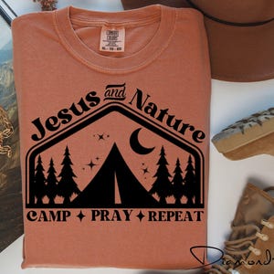 May include: A rust-colored t-shirt with a black graphic design of a tent, trees, and a crescent moon. The text "Jesus and Nature" is above the graphic and "Camp Pray Repeat" is below the graphic.