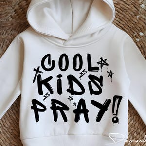May include: A white hooded sweatshirt with the text "COOL KIDS PRAY!" in black graffiti-style lettering. The design includes stars and lightning bolt accents. The sweatshirt is a casual style.
