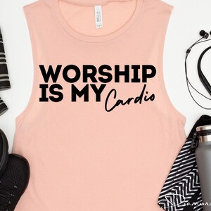 May include: A pink sleeveless tank top with the text "Worship is my Cardio" in black lettering. The tank top is on a white surface with other fitness accessories.