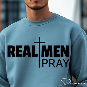 May include: A light blue sweatshirt with a black cross and the text "REAL MEN PRAY" printed on the front.
