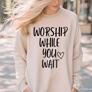 May include: Cream-colored sweatshirt with the text "WORSHIP WHILE YOU WAIT" in black script, with a heart symbol. The long-sleeved top is a casual style, suitable for everyday wear. The image is taken outdoors.