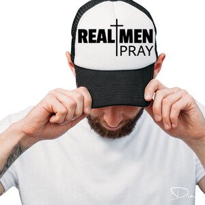 May include: A black and white trucker hat with a white front panel and a black mesh back. The front panel has the text "REAL MEN PRAY" in black letters with a cross between the words "REAL" and "MEN".