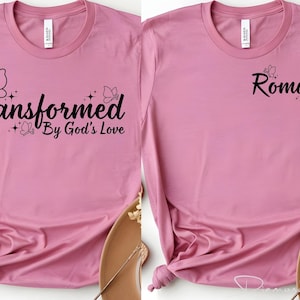Può includere: Due magliette rosa con testo nero e grafiche a farfalla. Una maglietta dice "Trasformato dall'amore di Dio" e l'altra dice "Romani 12:2". Le magliette sono esposte su una superficie marrone chiaro.