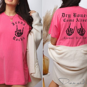 May include: Pink t-shirt with a black graphic of a skeleton hand making a rock and roll sign. The text "Jesus Rocks" is printed below the hand. The back of the shirt has a black graphic of two skeleton hands making a rock and roll sign with the text "Dry Bones Come Alive (Ezekiel 37:1-10)".