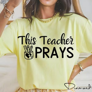 May include: A yellow t-shirt with the text "This Teacher Prays" in black. A black and white illustration of a pencil holder with pencils and a heart with a cross inside is above the text.