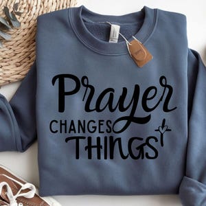 May include: A blue sweatshirt with the text "Prayer Changes Things" in black lettering. A small cross with a heart is next to the text.