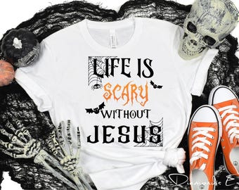 以下が含まれることがあります： 「LIFE IS SCARY WITHOUT JESUS」という黒とオレンジの文字と、クモの巣、クモ、2匹のコウモリのグラフィックがプリントされた白いTシャツ。Tシャツは、頭蓋骨、小さなカボチャ、オレンジ色のスニーカーと一緒に配置されています。
