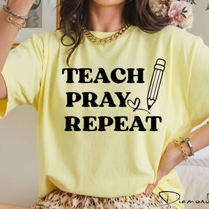 May include: A light yellow short-sleeved t-shirt featuring black text that reads TEACH PRAY REPEAT. The design includes a graphic of a pencil next to the word PRAY and a small heart symbol. The shirt has a crew neckline and a standard fit.