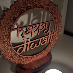 Pode incluir: Um letreiro de madeira com um design de mandala e as palavras "Happy Diwali" gravadas nele. O letreiro está sobre uma base de madeira e há uma vela acesa em primeiro plano.