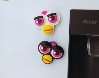 May include: Two whimsical, cartoon-style magnets featuring a face with large pink eyes, black eyelashes, and a yellow beak. One magnet has a white outline, while the other has a black outline. The magnets are affixed to a white surface.