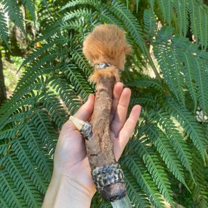 May include: A decorative wand featuring a textured brown wooden branch, topped with a fluffy tan fur pom-pom. The base of the wand is adorned with dark, overlapping scales and terminates in a clear, pointed crystal tip extending from the bottom.