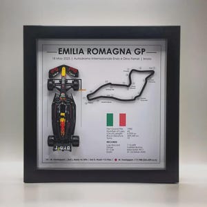Könnte beinhalten: Gerahmtes Kunstwerk mit einem Formel-1-Rennwagenmodell, dem Streckenlayout des Emilia Romagna GP und Renndetails. Der schwarze Rahmen umgibt einen weißen Hintergrund mit Text und Grafiken. Das Auto ist rot, gelb und schwarz.