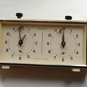 May include: Vintage chess clock with a cream-colored face and black hands. The clock has two dials, each with a black knob on top. The text "lantaa" and "MADE IN USSR" are visible on the clock face.