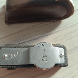 May include: A vintage light meter with a brown leather case. The meter has a silver dial with numbers and a logo. The word "Смена" is printed on the meter.
