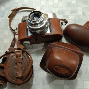 May include: A vintage silver camera with a brown leather case and two additional brown leather cases. The camera has a silver lens with the word "Monitor" printed on it.