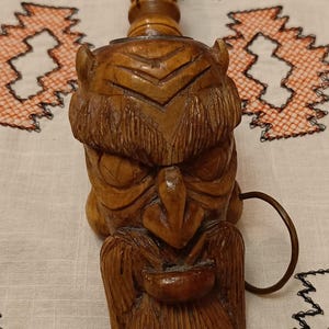 Puede incluir: Un tapón de botella de madera tallada a mano con una cara detallada y barba, más un anillo. El tapón es marrón y tiene una superficie texturizada. El fondo es un paño blanco con bordados naranjas.