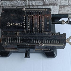 Puede incluir: Una calculadora mecánica negra vintage con el texto cirílico "Феликс" en la parte delantera. Cuenta con múltiples diales numéricos, palancas y una manivela. La calculadora está sobre una superficie blanca.