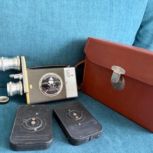 May include: A gray 16mm movie camera with two silver lenses, a circular dial with text, and a small lens cap. Included are two black rectangular film magazines and a brown leather carrying case with a silver metal latch closure.
