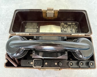 Pode incluir: Um telefone de campanha vintage de baquelite castanha numa caixa retangular. O dispositivo apresenta um auscultador preto no centro, um fecho metálico e diagramas técnicos internos com o texto "TAI-43" impresso na tampa interior.