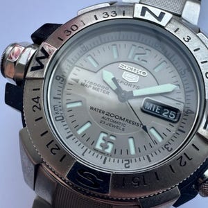 Puede incluir: Un reloj Seiko 5 Sports plateado con una correa de acero inoxidable. La esfera muestra la fecha, la hora y varias mediciones. El reloj es resistente al agua hasta 200 metros y tiene 23 joyas automáticas.