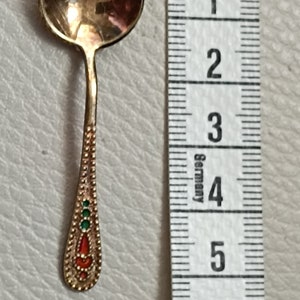 Könnte beinhalten: Ein kleiner, goldfarbener Löffel mit einer runden Schale und einem dekorativen Griff. Der Griff ist mit roten und grünen Emaille-Akzenten verziert. Der Löffel ist etwa 5 cm lang.
