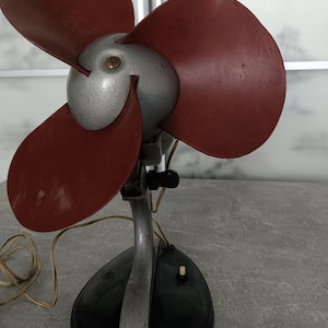 Könnte beinhalten: Ein elektrischer Tischventilator im Vintage-Stil mit drei dunkelroten Flügeln an einem silbernen Motorgehäuse. Der Ventilator wird von einem geschwungenen silbernen Metallständer auf einem schwarzen Sockel mit weißem Schalter und Kabel gehalten.